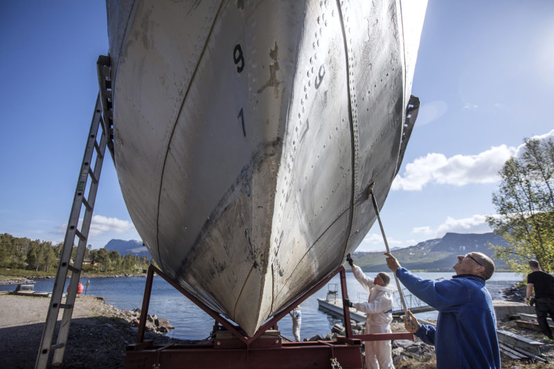 Två personer på dugnad står på marken och tvättar skrovet på en båt i docka. I bakgrunden syns sjö och fjäll.