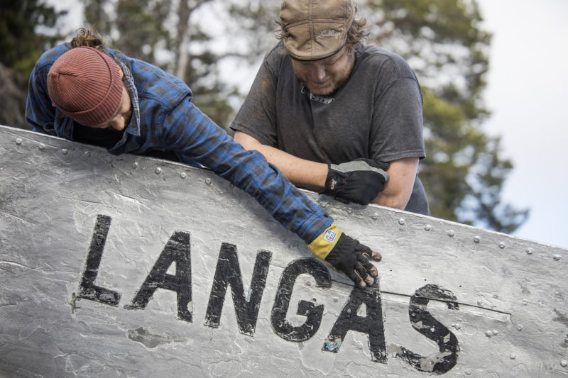 Två personer på dugnad böjer sig över relingen på en båt och granskar dess ditmålade namn: Langas.