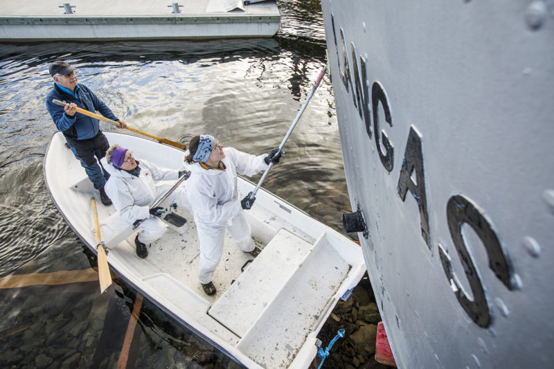 Tre personer står i en mindre båt och tvättar därifrån skrovet på ett större fartyg. Dugnad.