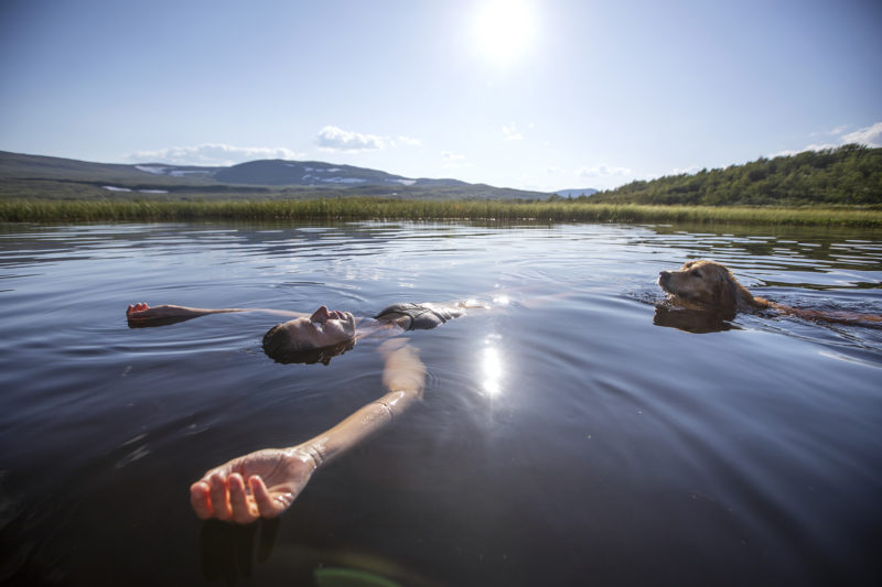 En kvinna och en hund flyter i vattenytan med ett fjäll i bakgrunden.