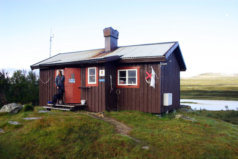 En kvinna och en hund i dörröppningen till en fjällstuga med sjö i bakgrunden. Vandra i Jämtland.