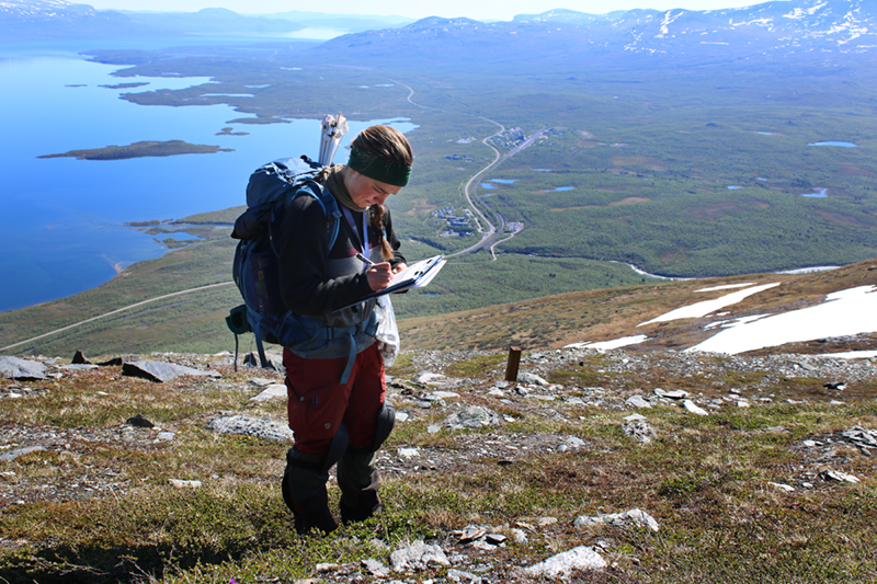 Kvinna med ryggsäck står på fjällsluttning med och läser dokument.