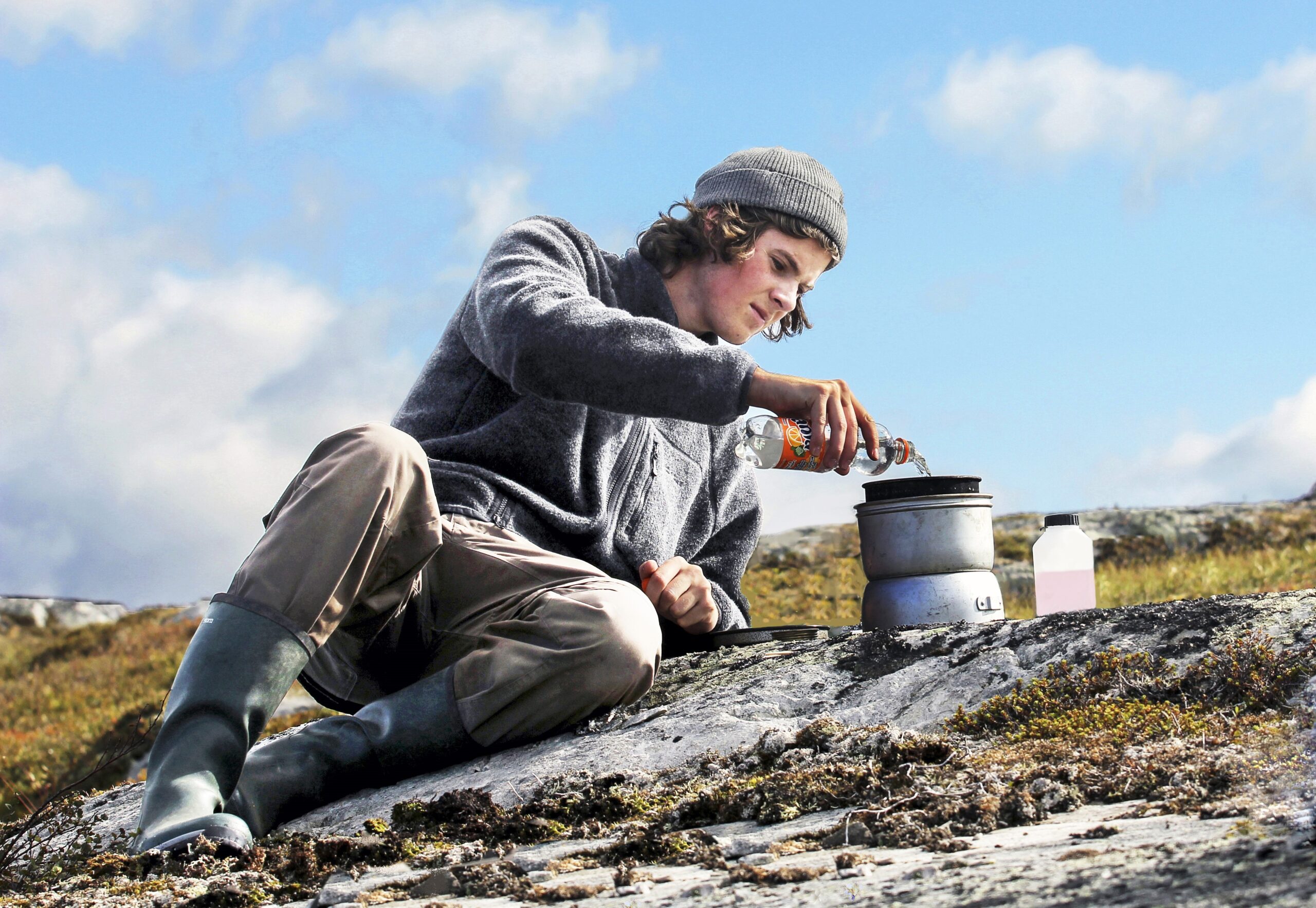 Laga mat utomhus. Ung man i gråa friluftskläder vilar mot en klippa i fjällmiljö och häller vatten i ett Trangiakök.