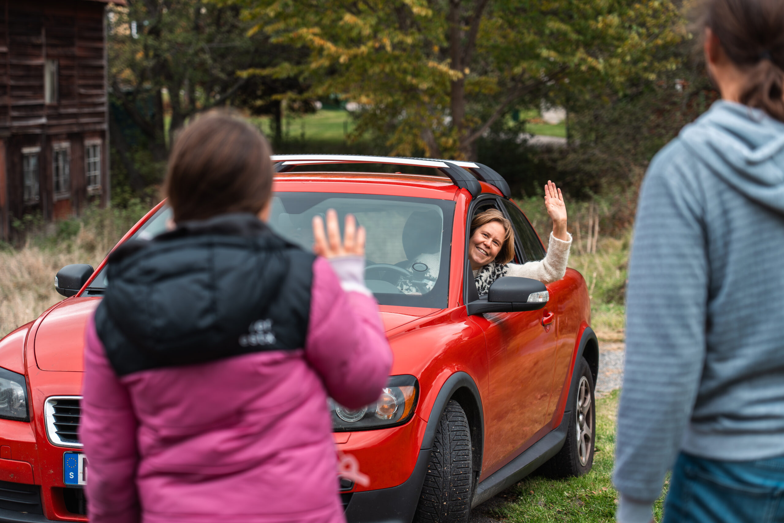 En kvinna i en röd bil hälsar glatt genom sidorutan på två personer som väntar på skjuts när Scouterna ansluter till Skjutsgruppen