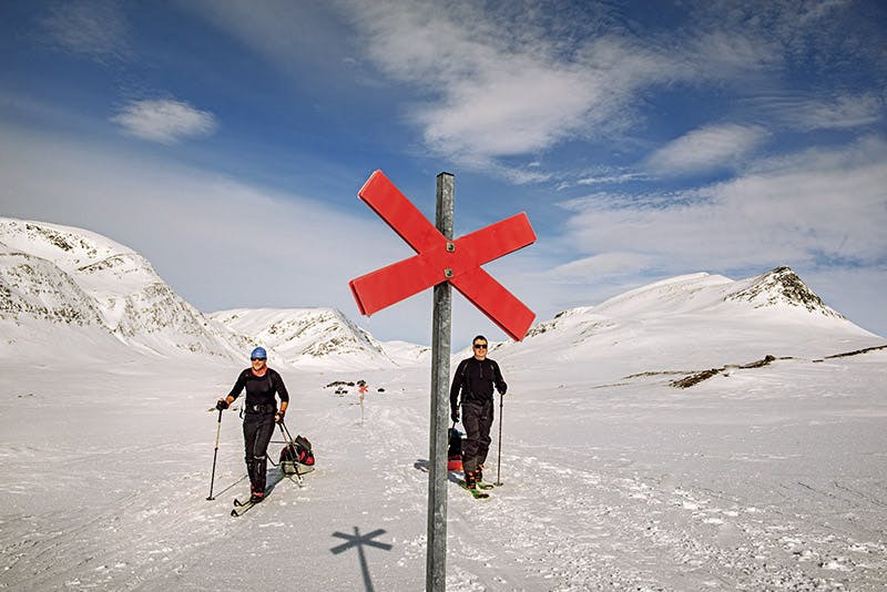 Två män åker skidor på var sida om ett ledkryss i de snötäckta Kirunafjällen.