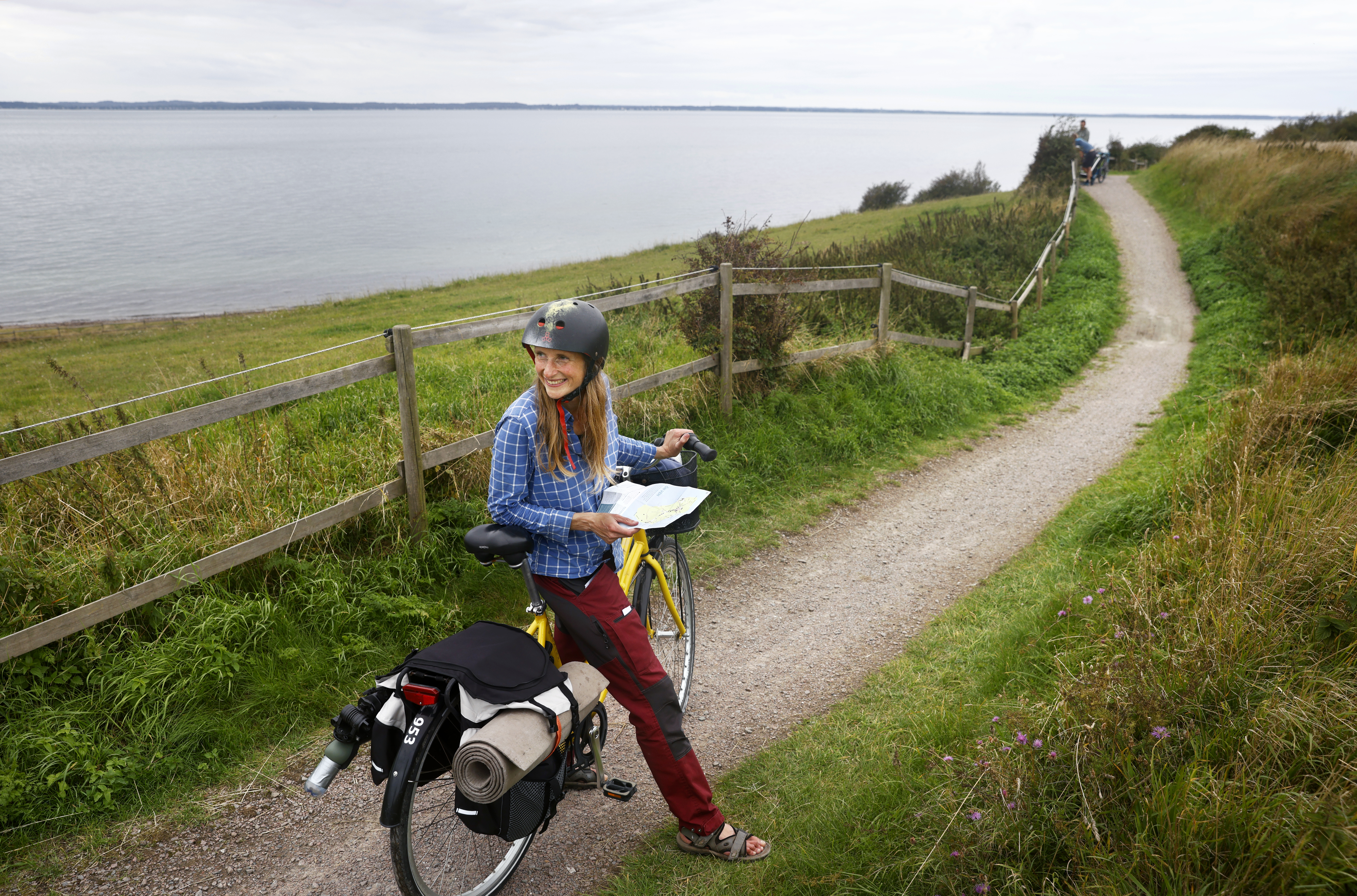 En kvinna i rutig skjorta har stannat med sin cykel på en grusväg vid vattnet.