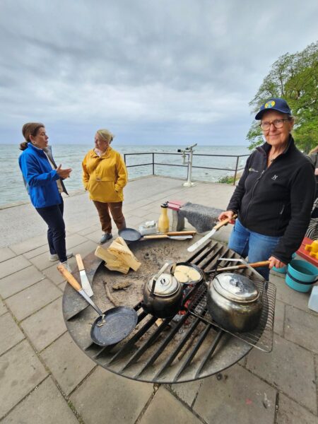 En kvinna grillar över öppen eld, två kvinnor talar med varandra i bakgrunden i Almedalen.
