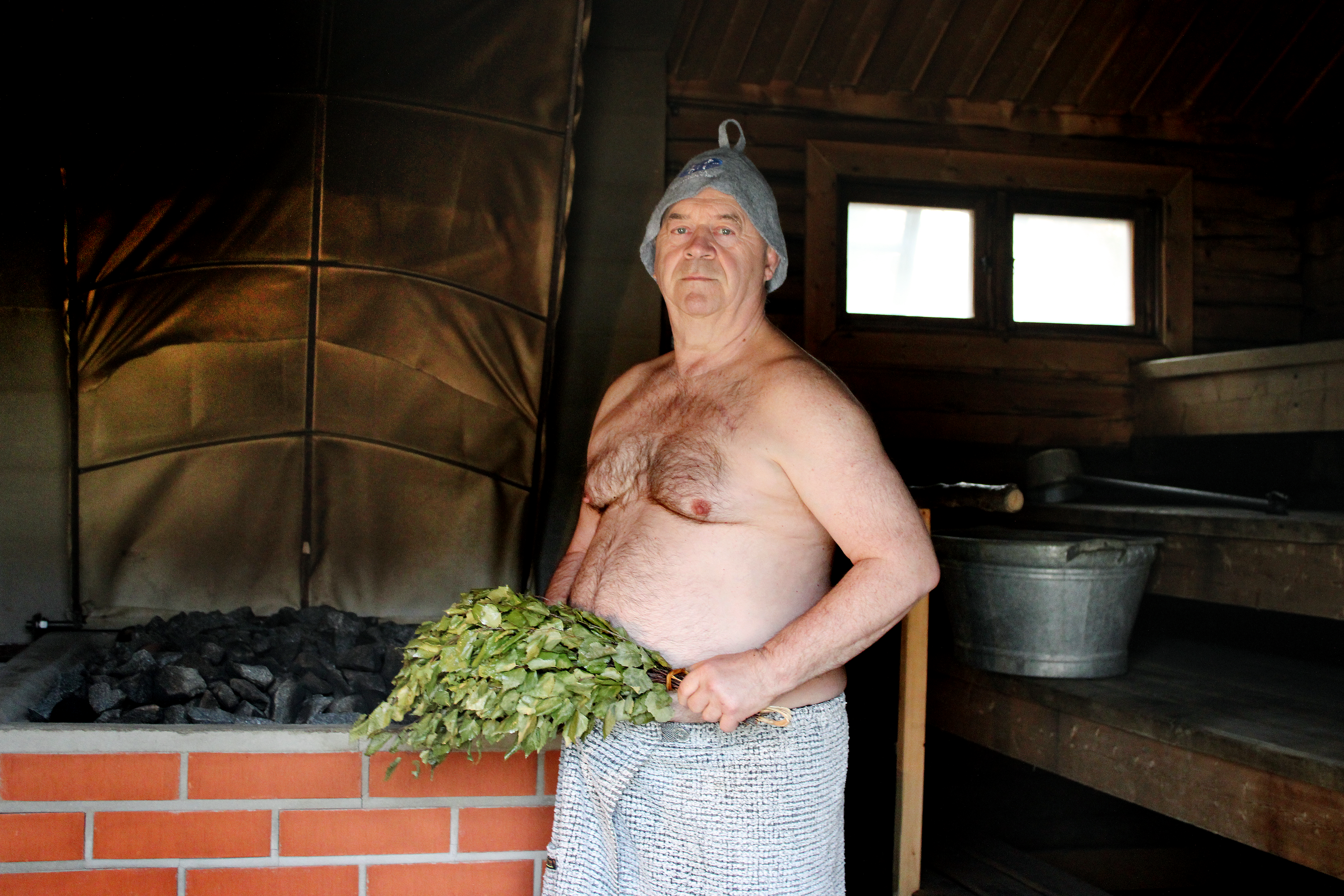 En äldre man med bar överkropp iklädd handduk och bastumössa med en björklövsruska i handen. Ordförande i Svenska Bastuakademien.