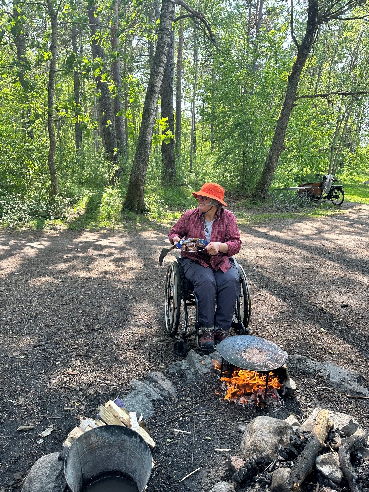 Kvinna i hatt och i rullstol sitter framför eldstad och grillar bröd.