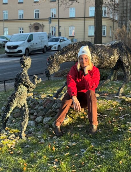 Turists chefredaktör klädd i röd anorak och vit mössa framför en bronsstaty föreställande en pojke och en häst.