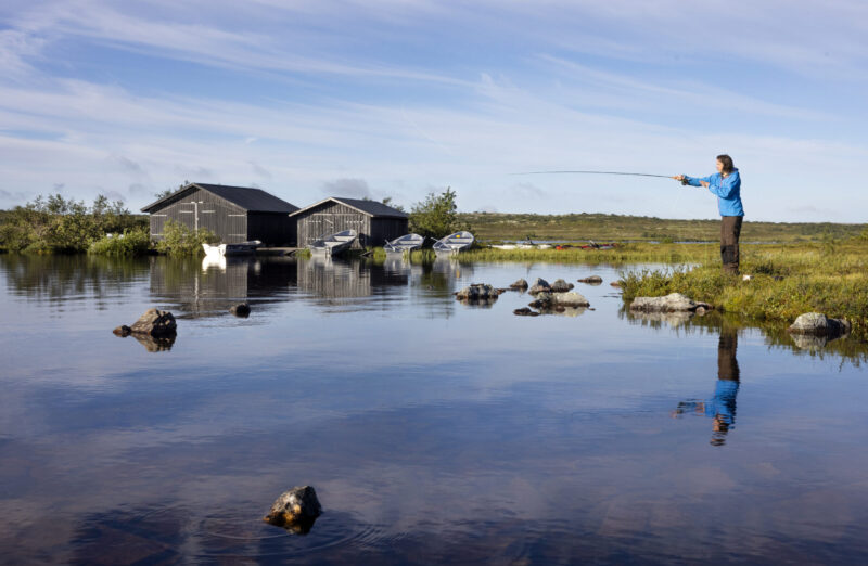 Person i blå jacka kastar med fiskespö över en spegelblank sjö med två grånade sjöbodar i bakgrunden.