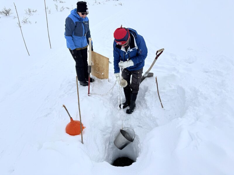 Två personer i blåa jackor hissar upp en hink med vatten ur ett hål i snön.