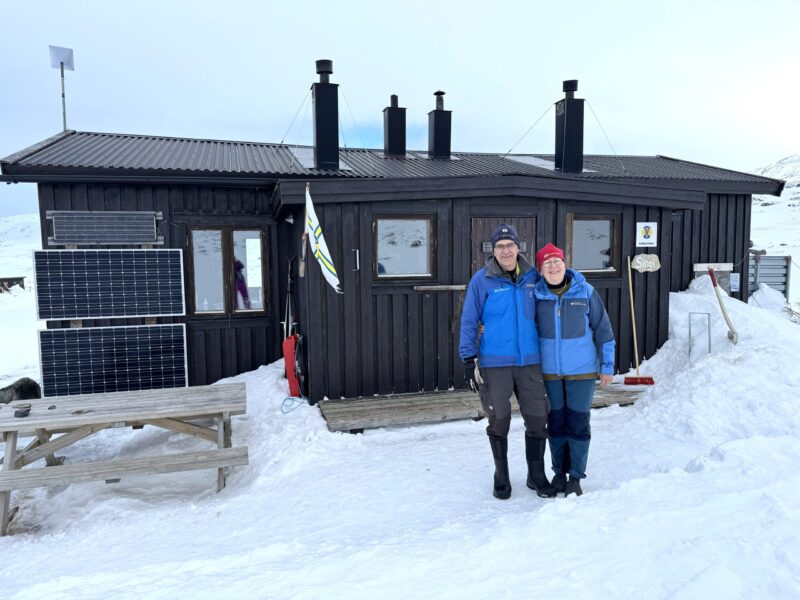Två personer i blåa jackor framför en fjällstuga.