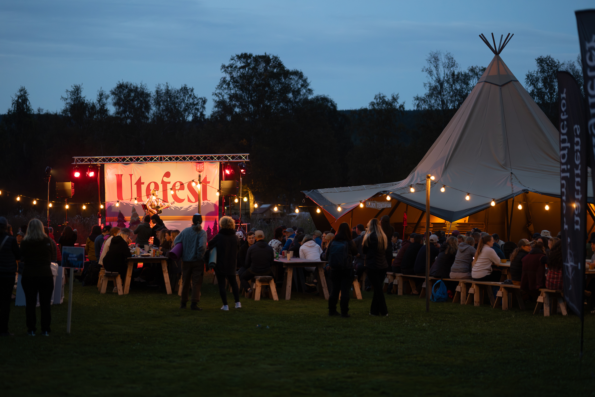 Kvällsbild av människor på friluftsfestival. Ljusslingor, en tältkåta och bord och bänkar samt en person som spelar musik från en scen.