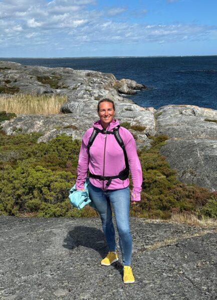 Kvinna i lila luvtröja, blå jeans och gula sneakers på en klippa med havet i bakgrunden.