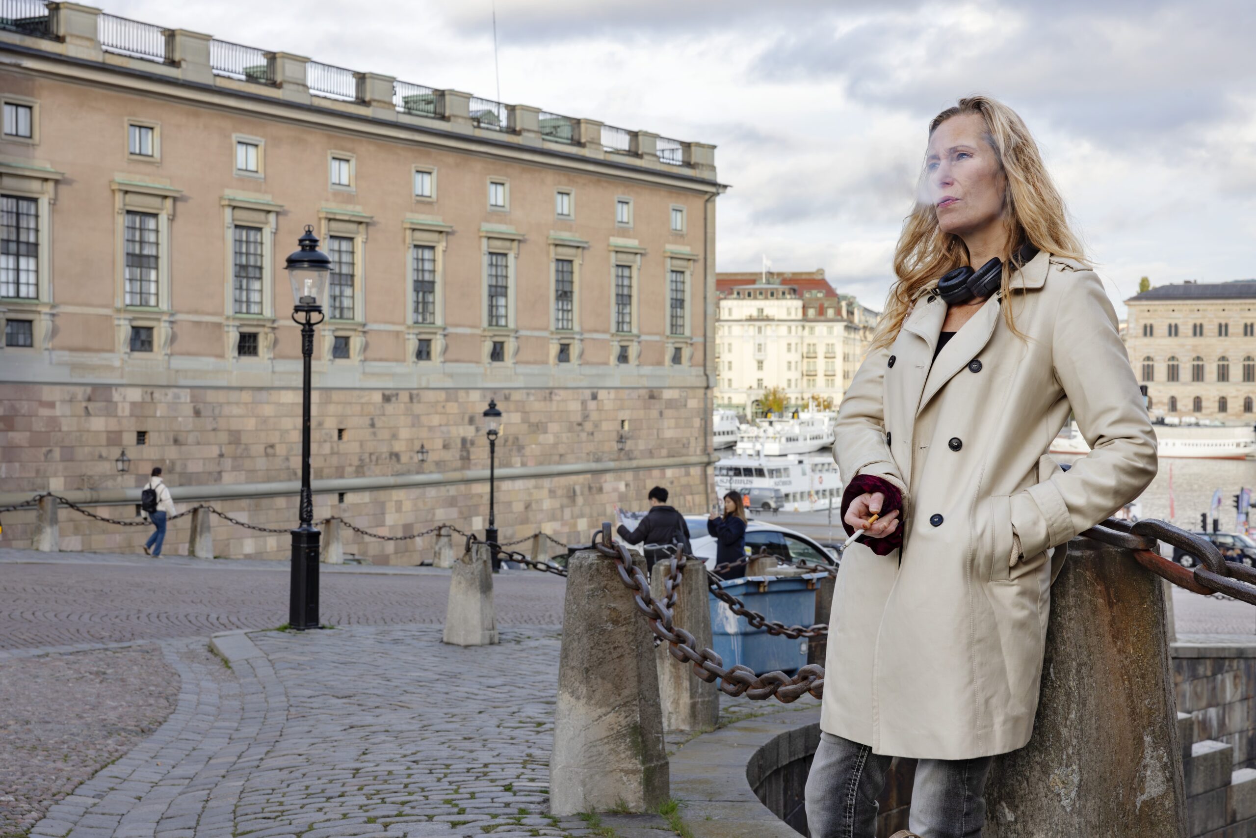 Kvinna i trenchcoat lutar sig mot en pelare med Stockholms slott i bakgrunden under en vandring om att leva som hemlös.