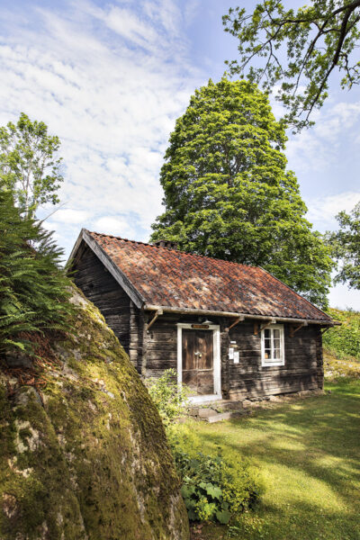 En bergsknalle i förgrunden och en grå timmerstuga med tegeltak på hembygdsgården i Ödeshög.