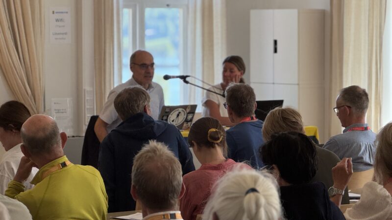 Bakhuvudena av flera människor i förgrunden och två personer på ett podium i bakgrunden.