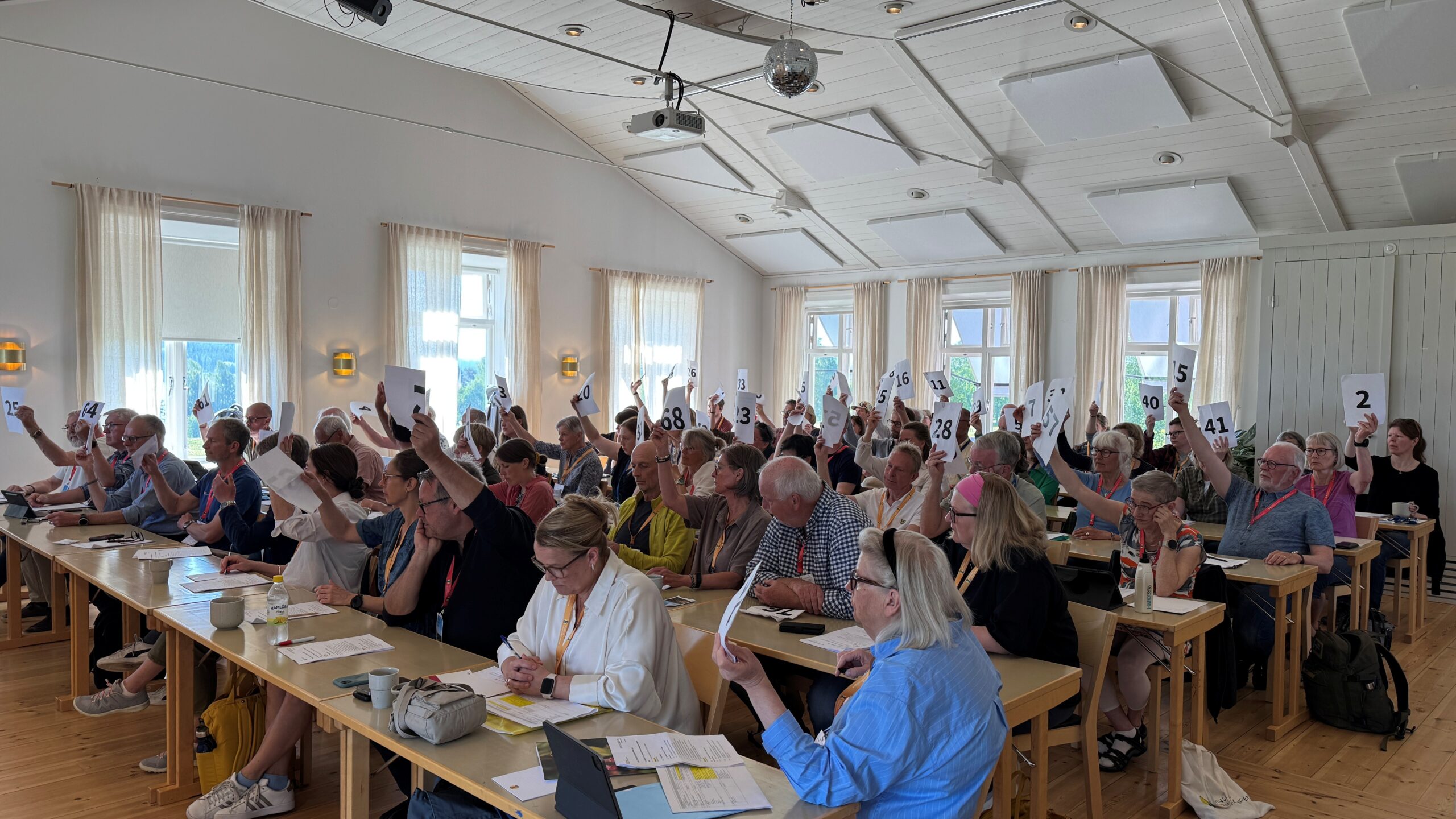Människor sitter längs bordsrader och håller upp lappar med nummer på under en omröstning.