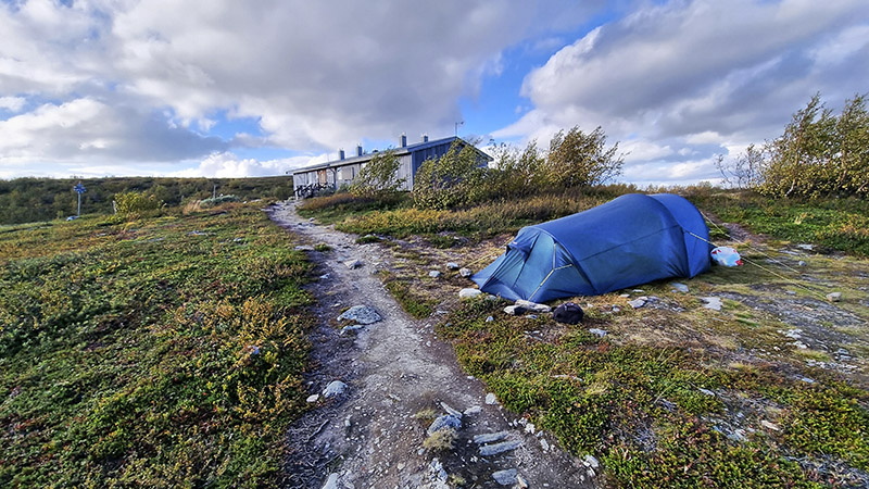 Ett blått tält på en stenig fjällbacke. I bakgrunden en grå stuga på Kungsleden.