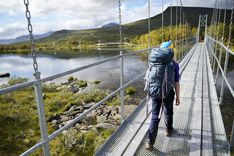 Ett barn med blå ryggsäck går på en hängbro i metall över ett vattendrag med fjälltoppar i bakgrunden.
