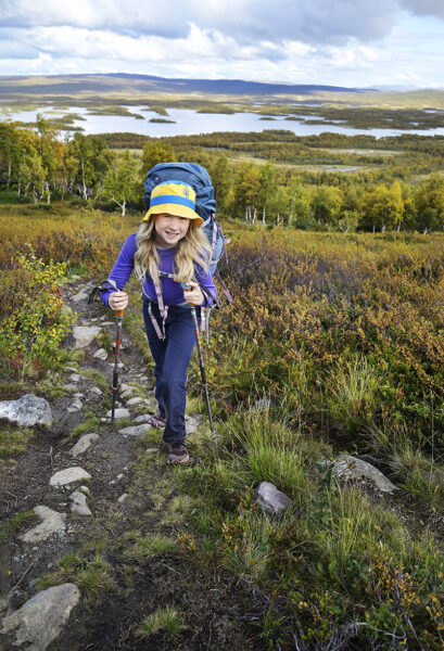 En flicka i lila tröja och blågul solhatt stöder sig på vandringsstavar när hon går uppför en stenig stig. I bakgrunden fjällbjörkskog och en sjö längs Kungsleden.