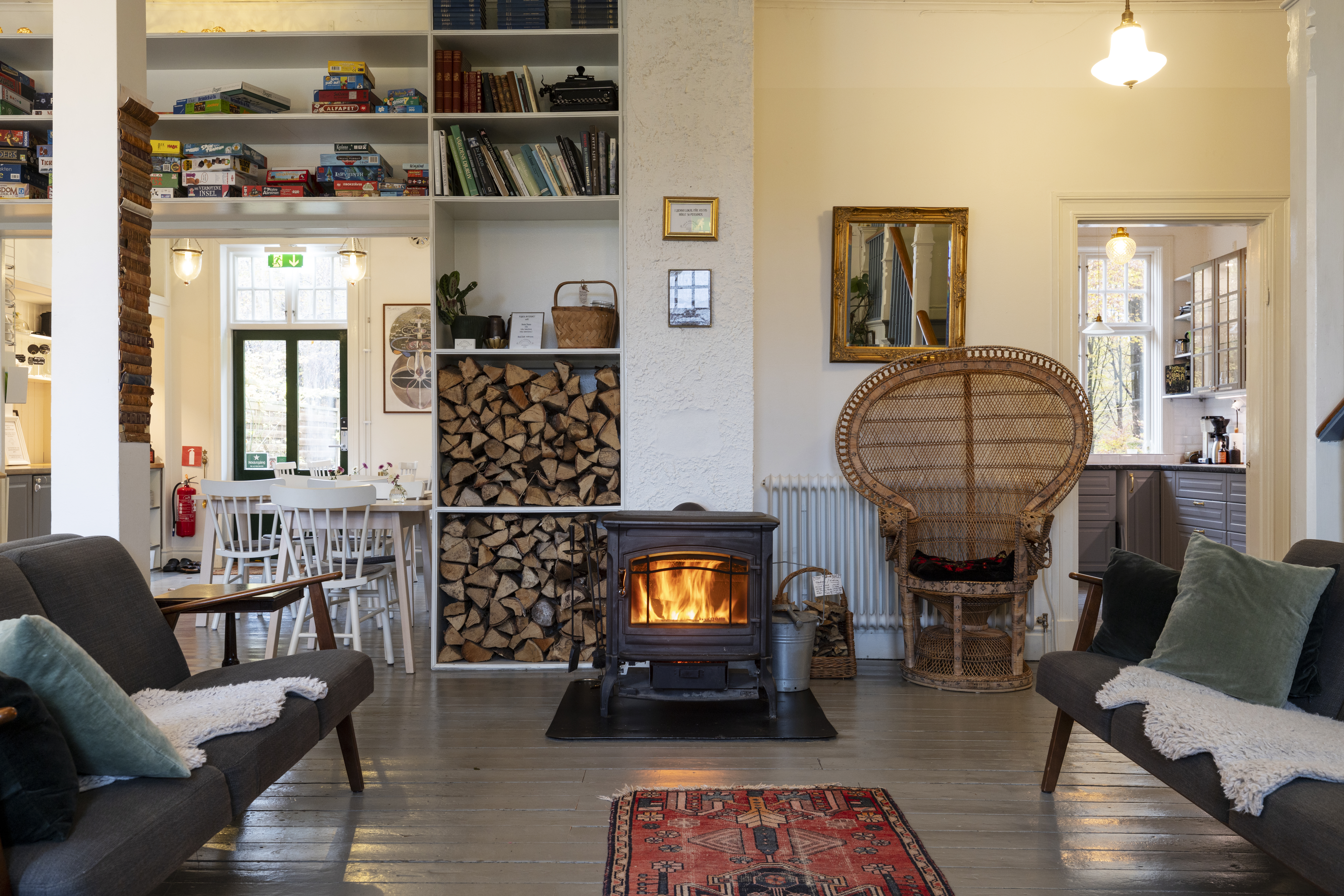 Ett rum med en sprakande brasa i en kamin i mitten. Till vänster en soffa och en platsbyggd bokhylla med spel och ved. Till höger en korgstol och ännu en soffa.