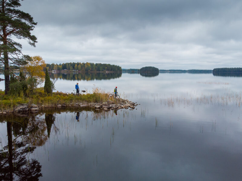 En spegelblank sjö. Till vänster i bild en udde där två cyklister ses stå och blicka ut över vattnet.