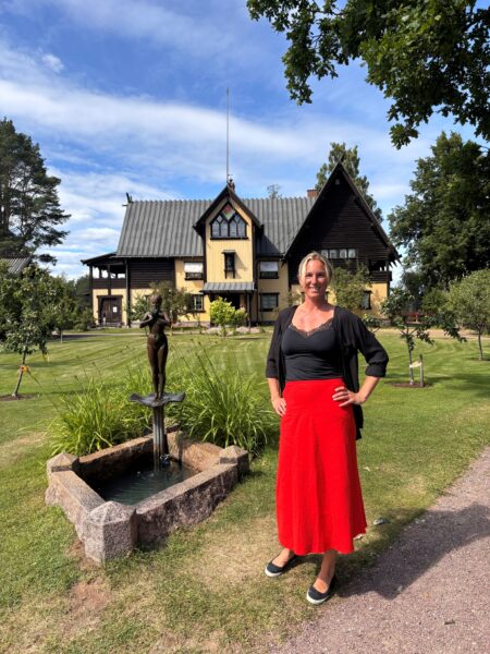 En kvinna i röd kjol och svart blus står bredvid en bronsstaty framför en trävilla som tillhört Anders och Emma Zorn.