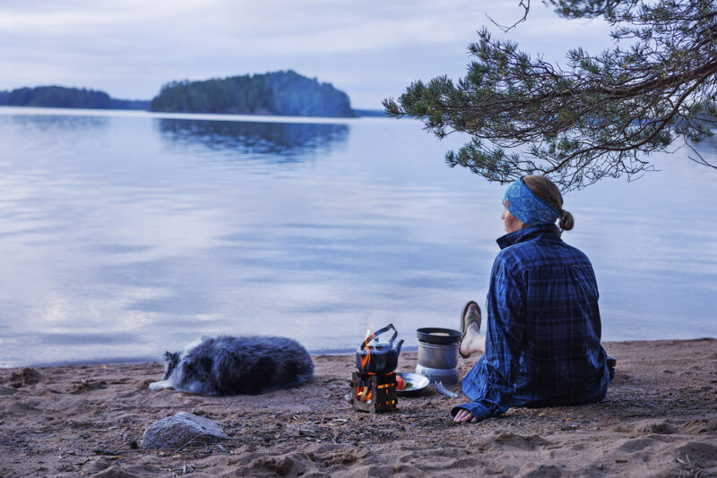 En kvinna i blåa kläder sitter på en sandstrand och tittar ut över ett stilla vatten. En kaffekanna står över en eld och en hund sover intill.