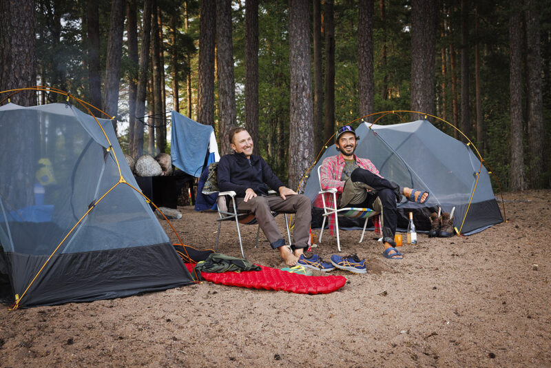 Två män sitter i campingstolar mellan två kupoltält på en sandstrand.